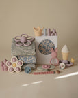 Mushie Extra Soft Muslin Crib Sheet displayed with assorted pastel baby toys on a light background.