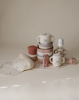 Mushie Bluey Gift Set: neutral silicone cups, plates, utensils, bib & bottle on a table.