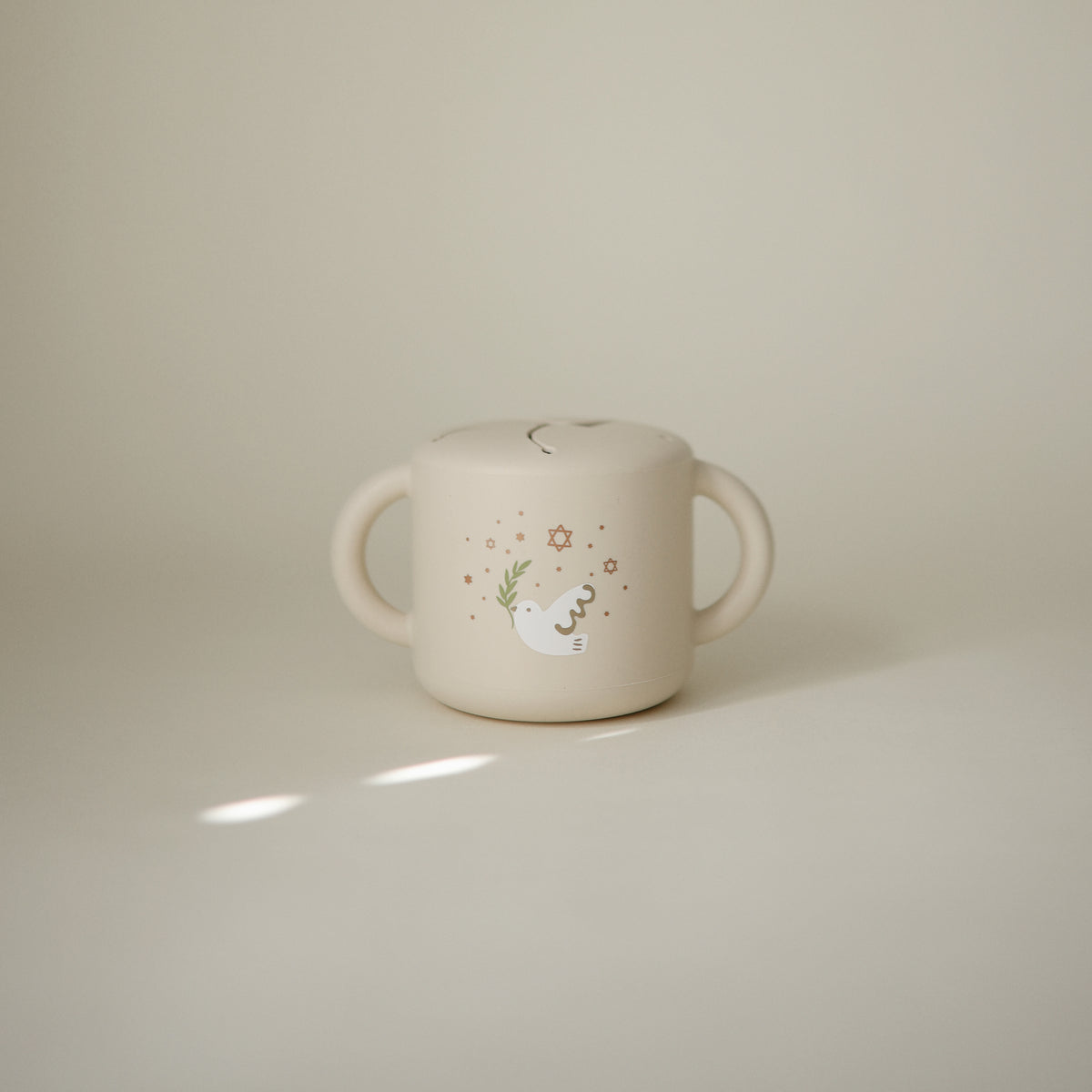 Mushie Snack Cup in beige, spill-proof with two handles and a cute bird illustration for toddlers.