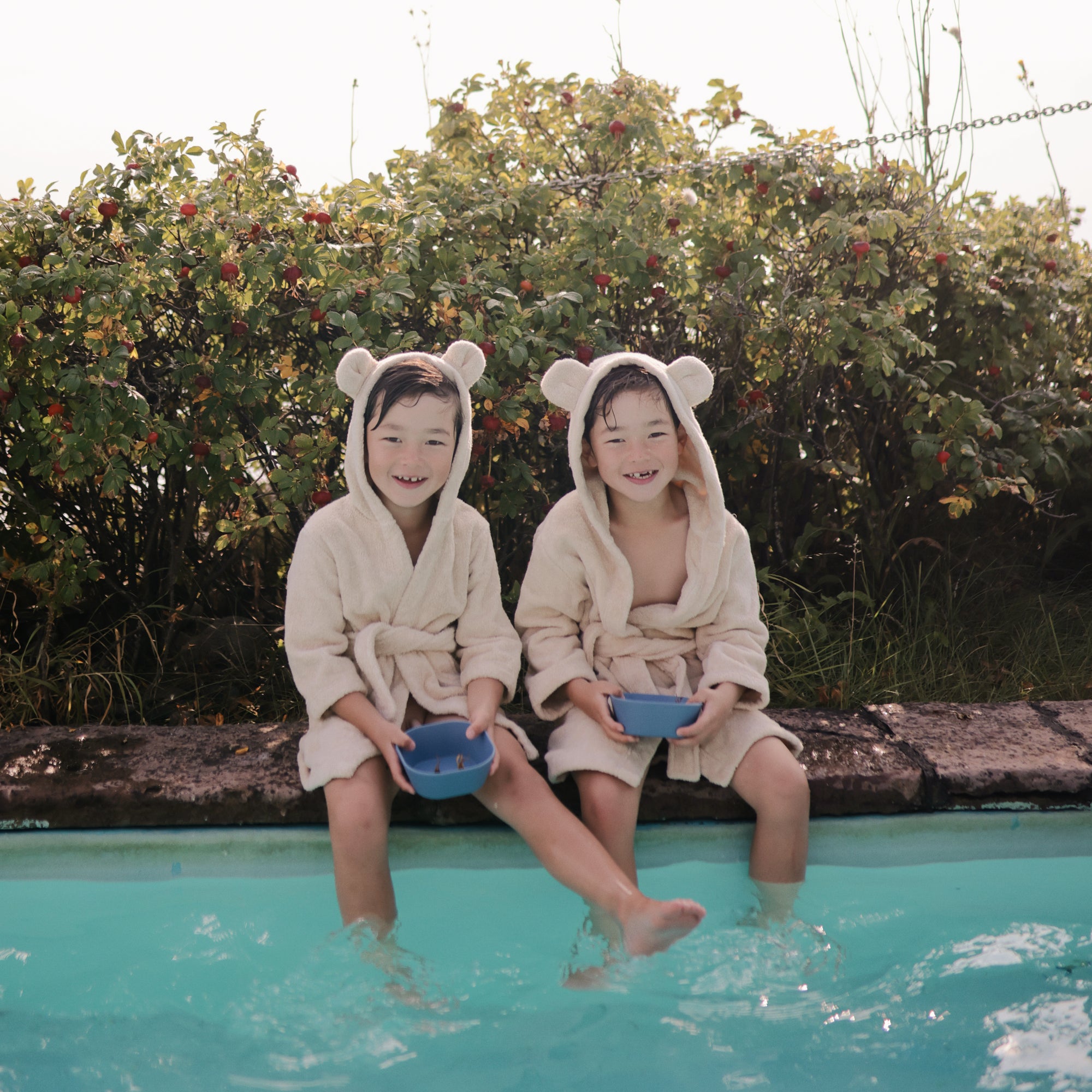 Two kids in bear ear robes by a pool hold mushie Square Dinnerware Bowls near green bushes.