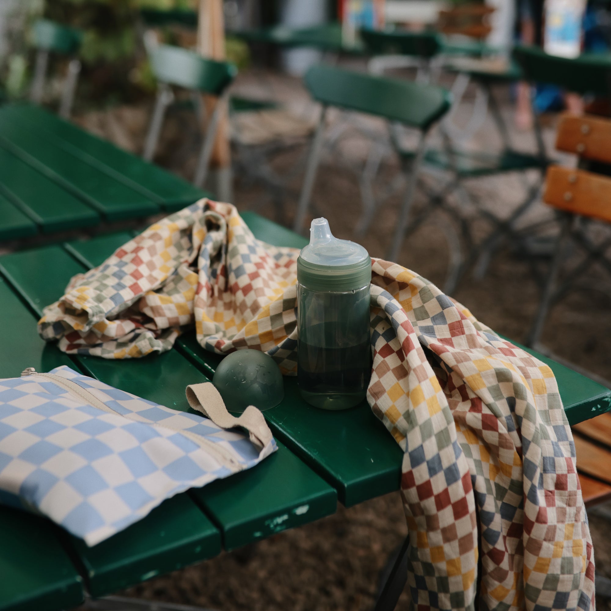 A sippy cup, mushie Organic Cotton Muslin Swaddle Blanket, and bag on a green outdoor table.