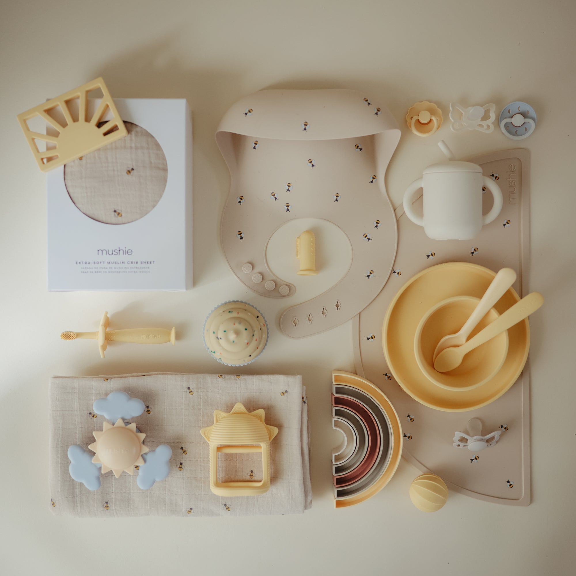 Extra Soft Muslin Crib Sheet by mushie displayed with pastel baby toys and feeding items.