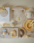 Extra Soft Muslin Crib Sheet by mushie displayed with pastel baby toys and feeding items.