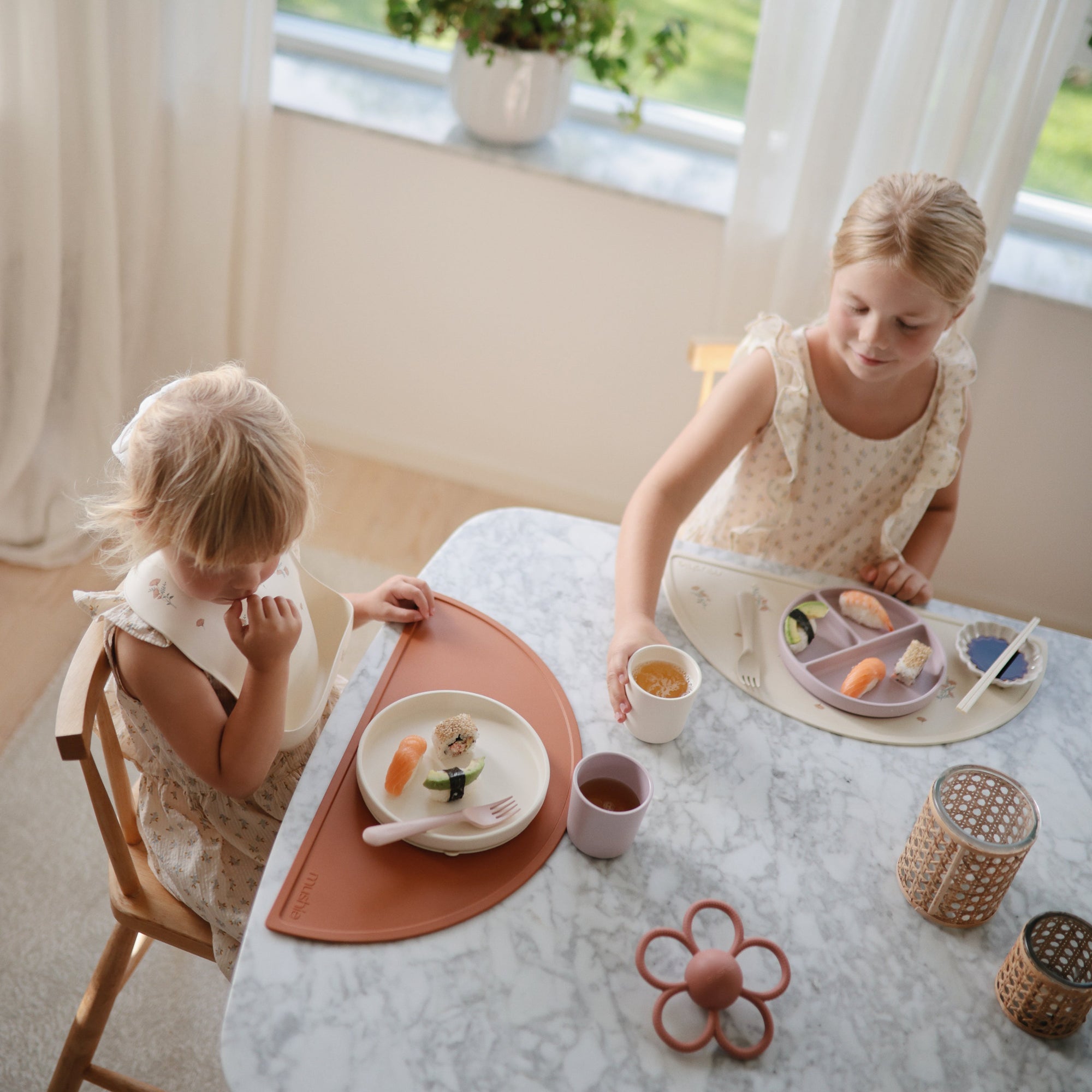 Two young children wear mushie Silicone Baby Bibs while eating at a marble table by a window.