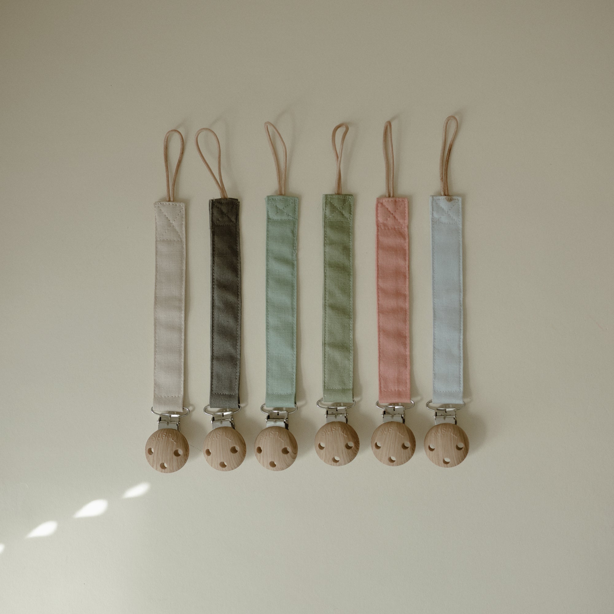 Two mushie linen pacifier clips with wooden clasps displayed on a light surface.