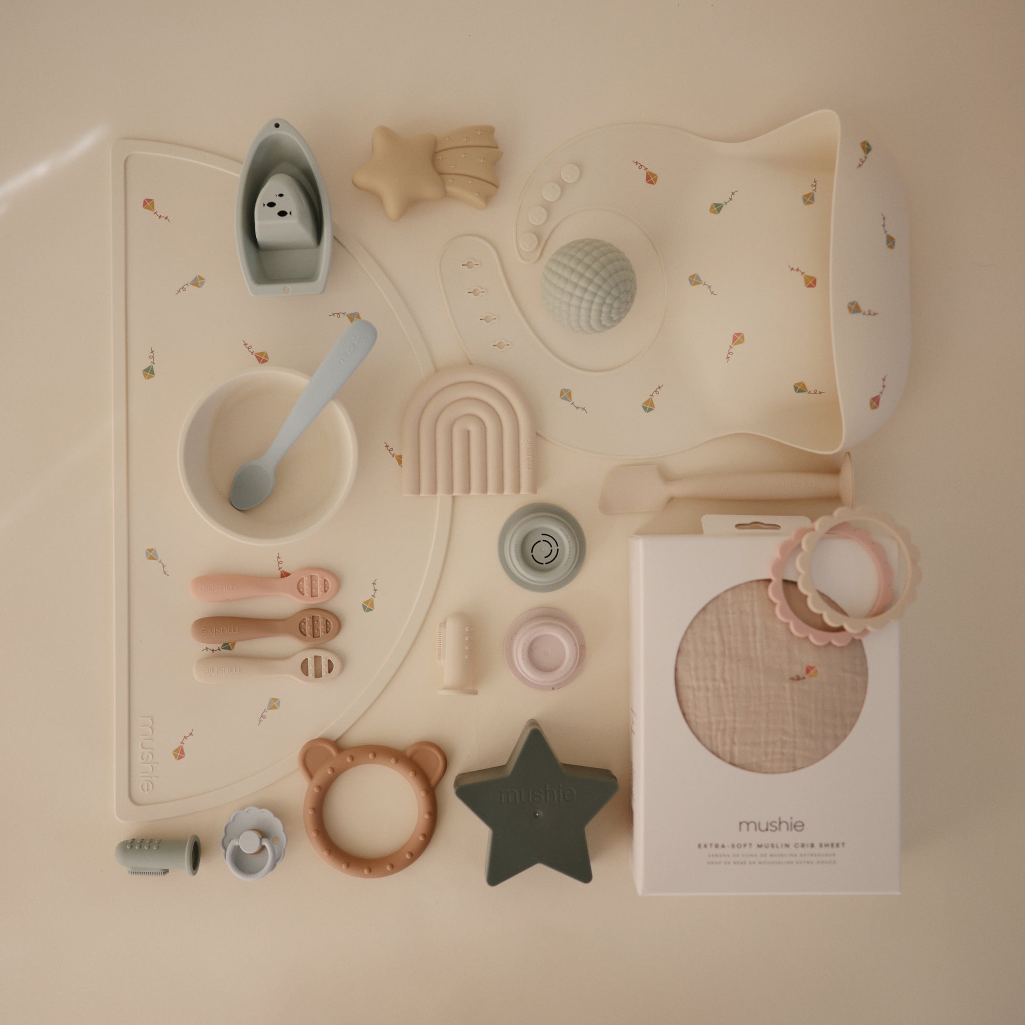 Mushie Extra Soft Muslin Crib Sheet displayed with assorted neutral baby feeding and teething items.