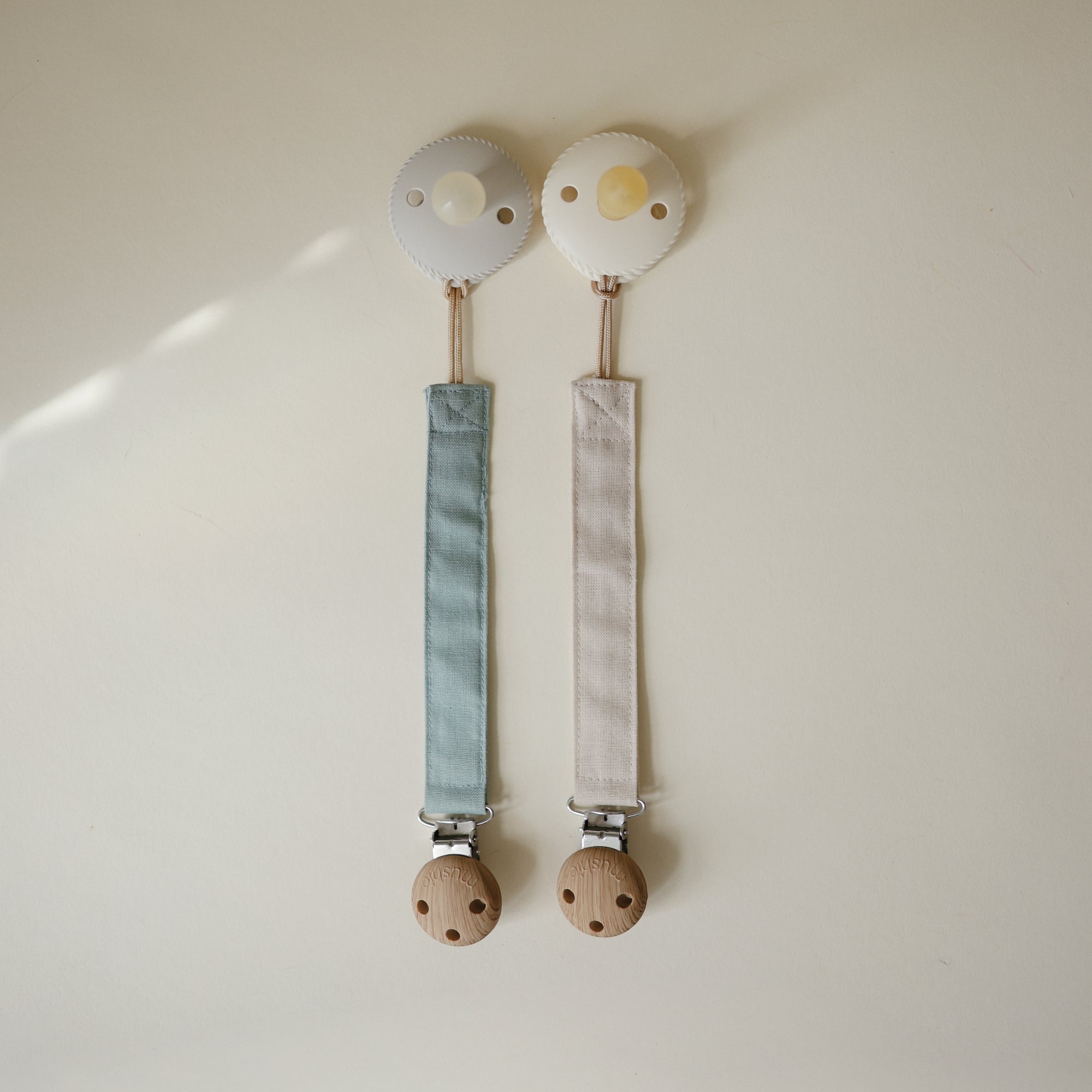 Mushie Linen Pacifier Clip 2-Pack: blue &amp; beige pacifiers with wooden clasps on light background.
