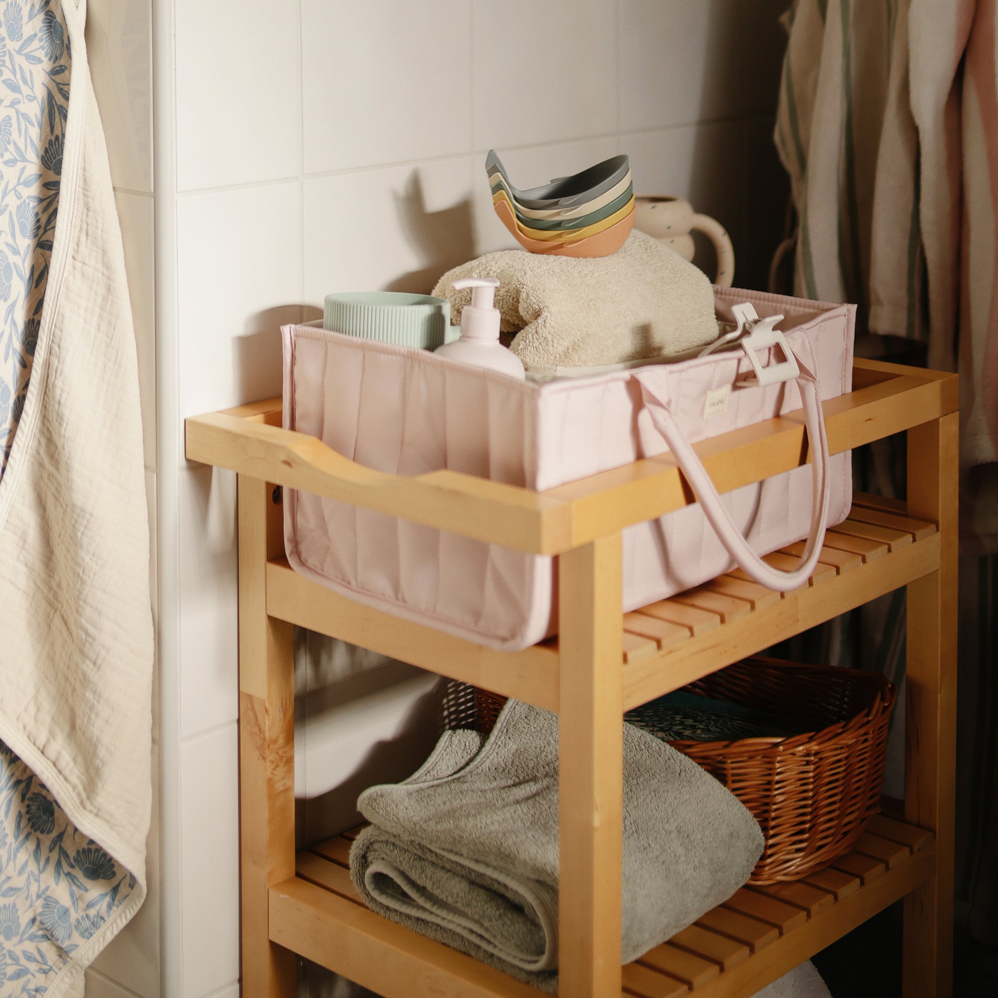 A mushie Diaper Caddy sits atop a wooden bathroom shelf with towels and toiletries.