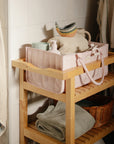 A mushie Diaper Caddy sits atop a wooden bathroom shelf with towels and toiletries.