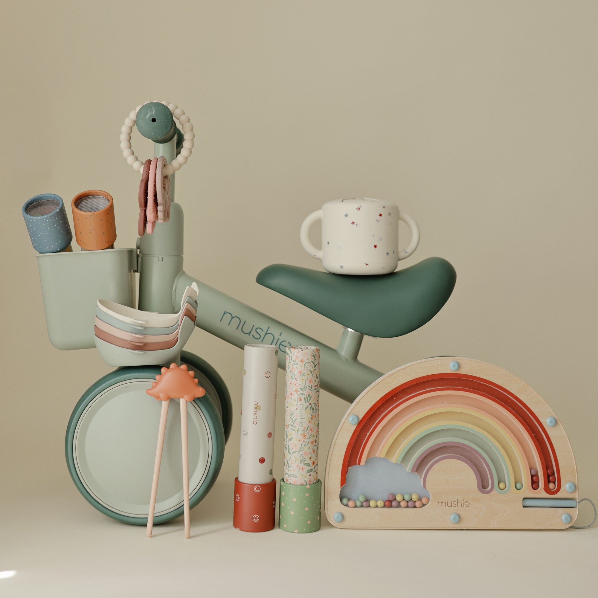 Mushie Mini Toddler Bike in green with pastel cups, bowls, and a wooden rainbow toy.