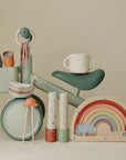 Mushie Mini Toddler Bike in green with pastel cups, bowls, and a wooden rainbow toy.