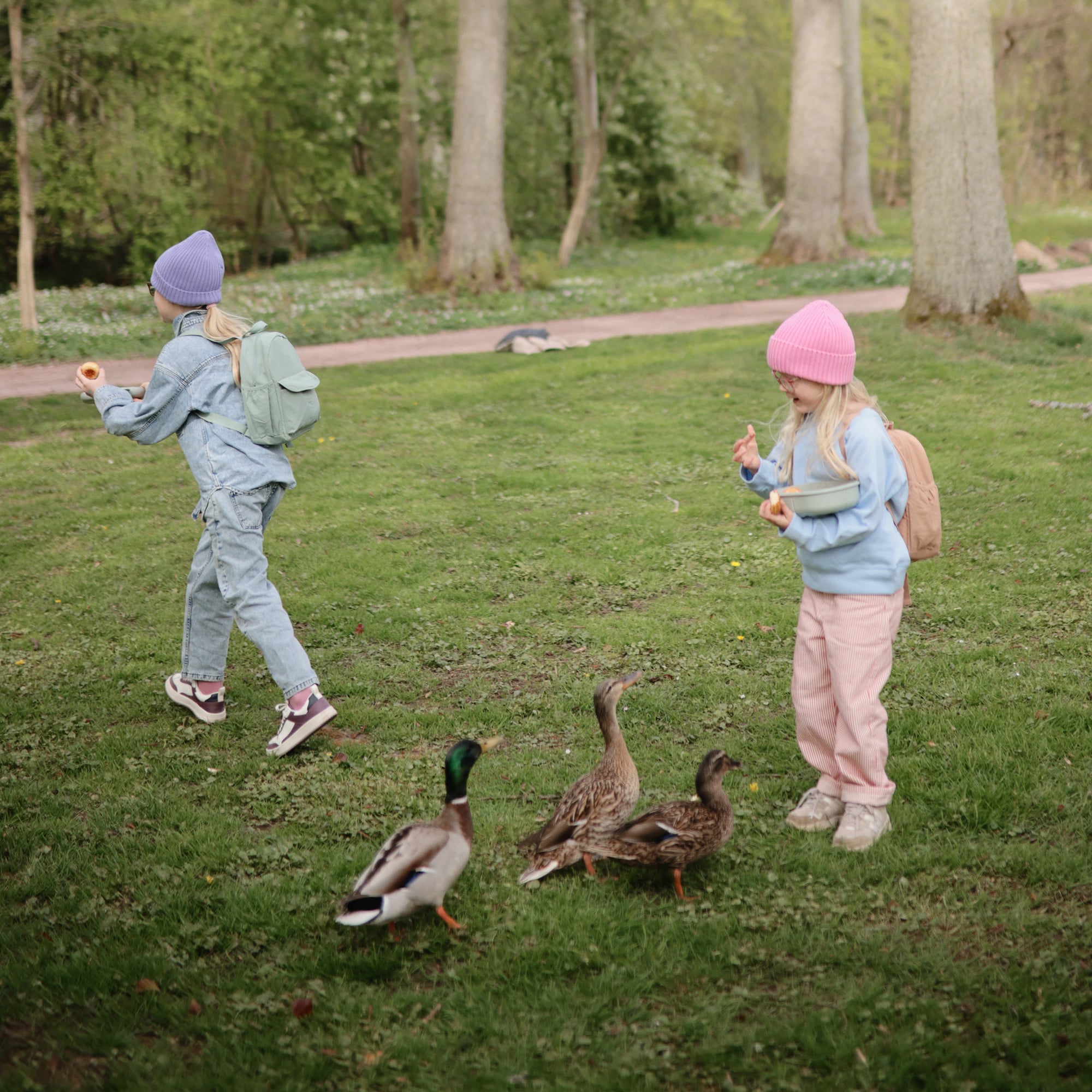 Two kids in pastel feed ducks, carrying mushie Lunchbox sets with matching fork and spoon.