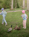 Two kids in pastel feed ducks, carrying mushie Lunchbox sets with matching fork and spoon.