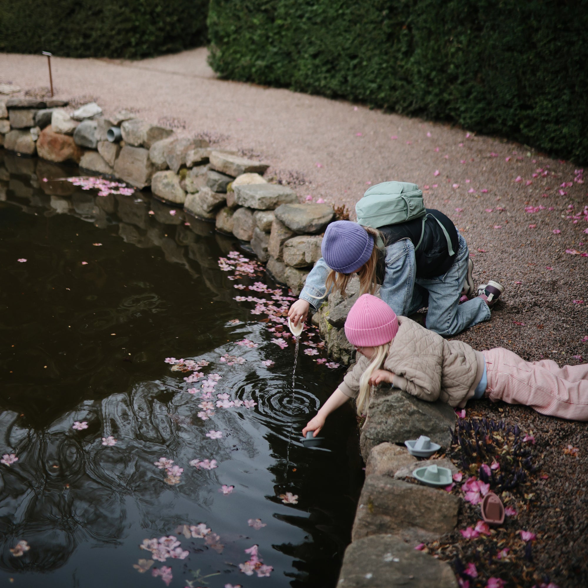 Two children play by a pond with mushie Bath Boats, wearing pastel hats and jackets.