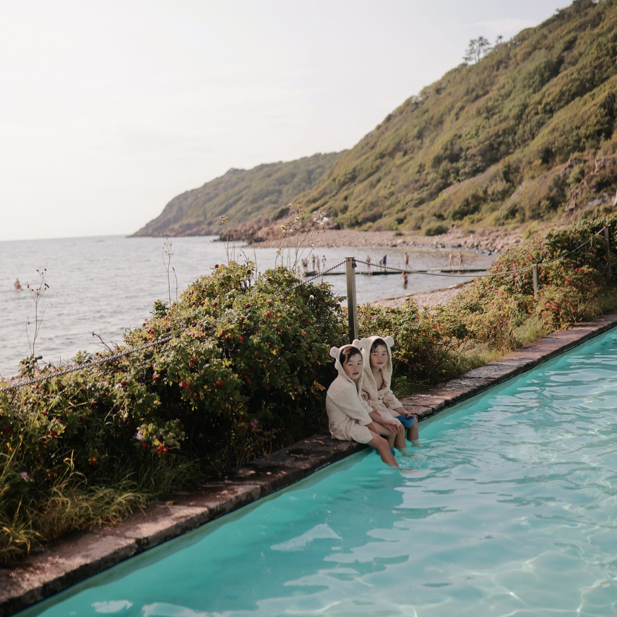Two kids in mushie Bear Robe sit by a pool with sea and hills in the background.