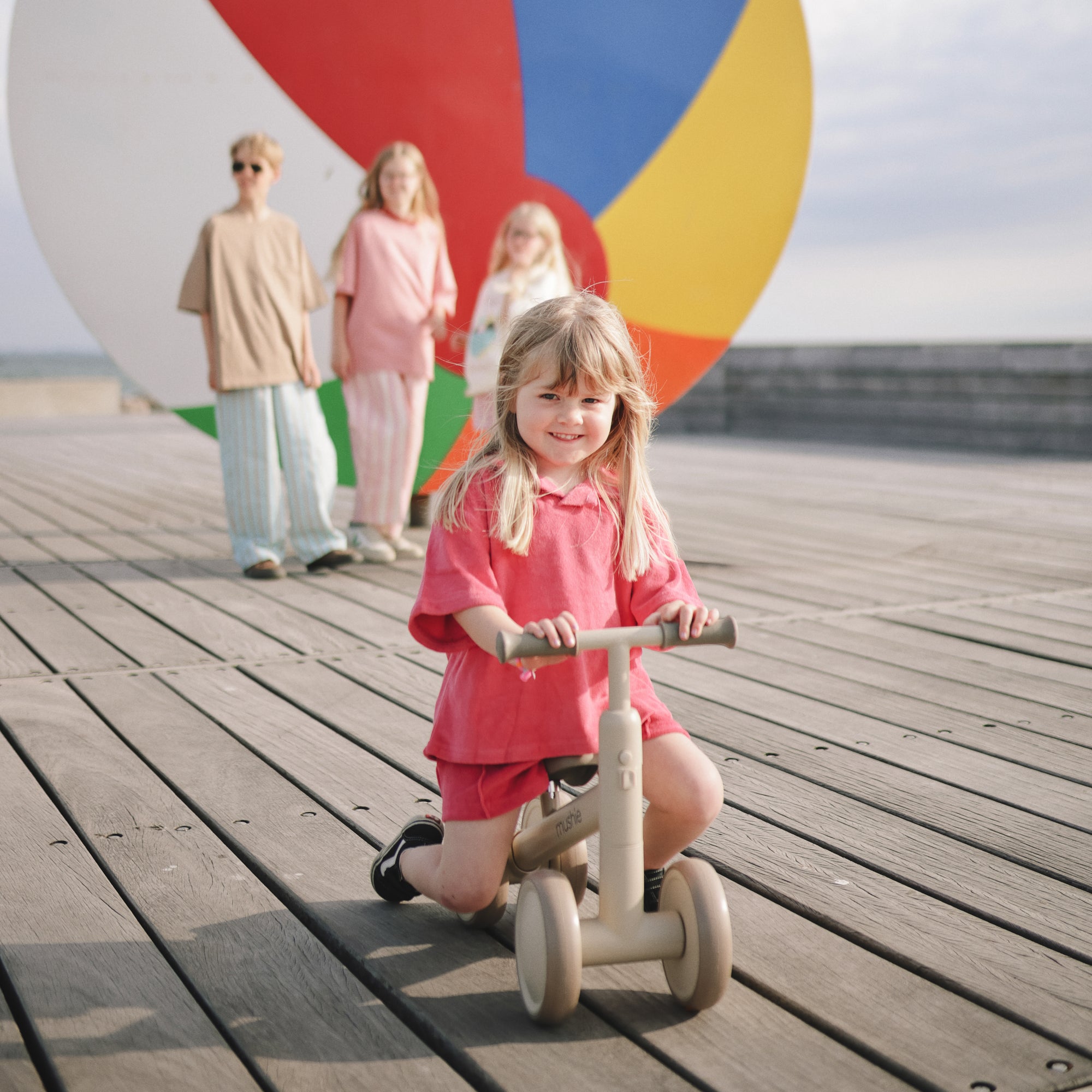 A young girl rides the mushie Mini Toddler Bike on a boardwalk, kids and beach ball behind her.