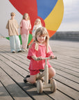 A young girl rides the mushie Mini Toddler Bike on a boardwalk, kids and beach ball behind her.