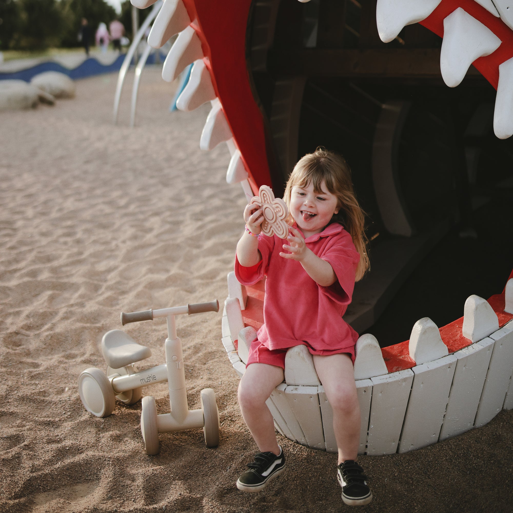 A young girl smiles happily on the playground beside her mushie Mini Toddler Bike.