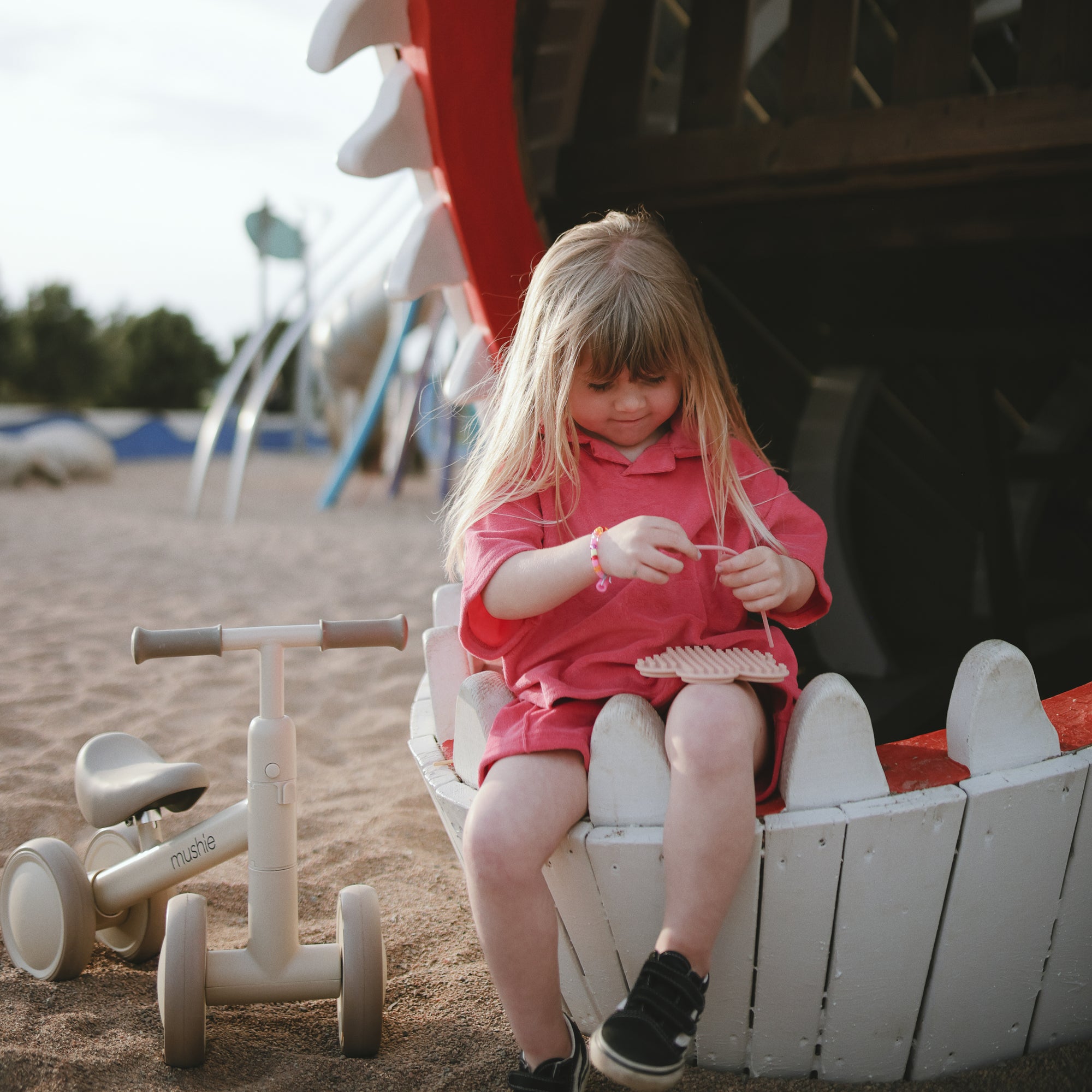 Young girl plays with mushie Silicone Sensory Toy, sitting near a white wooden structure.
