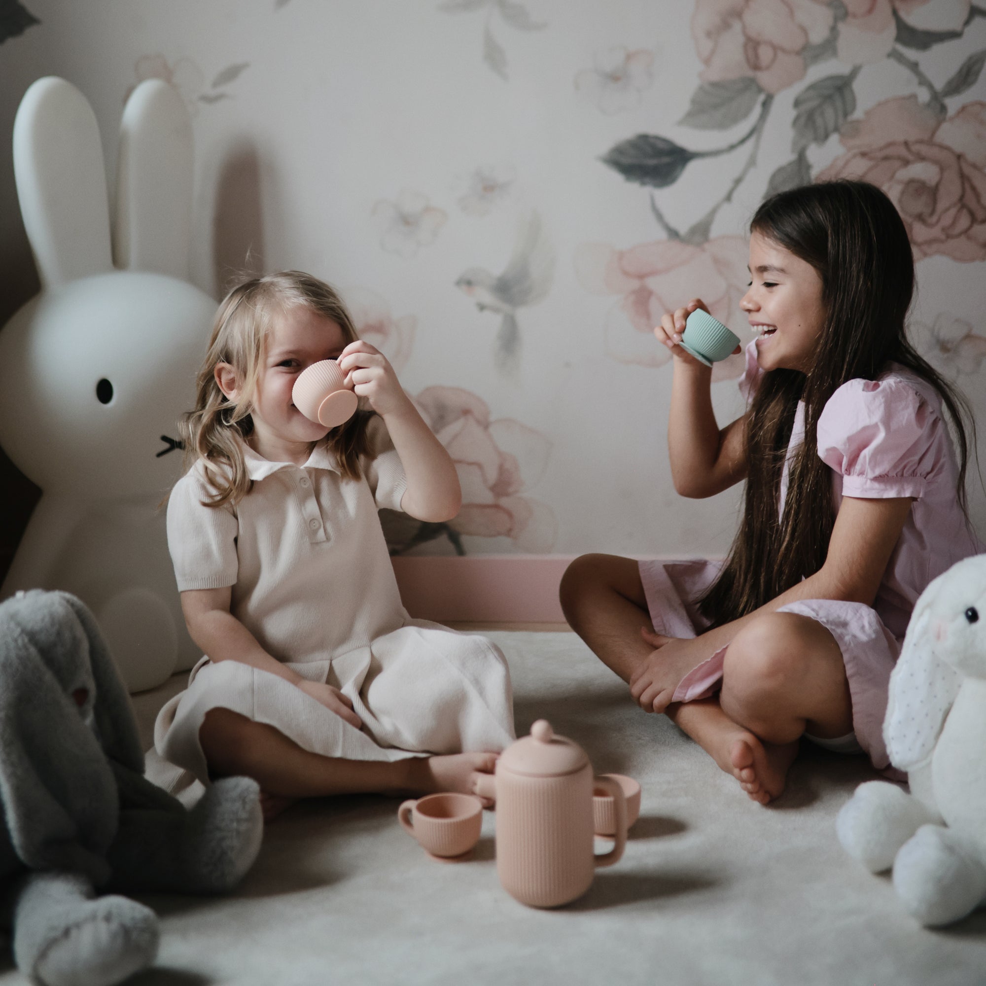 Two young girls enjoy the Mushie Silicone Tea Play Set in a softly lit, floral-themed room.
