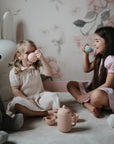 Two young girls enjoy the Mushie Silicone Tea Play Set in a softly lit, floral-themed room.