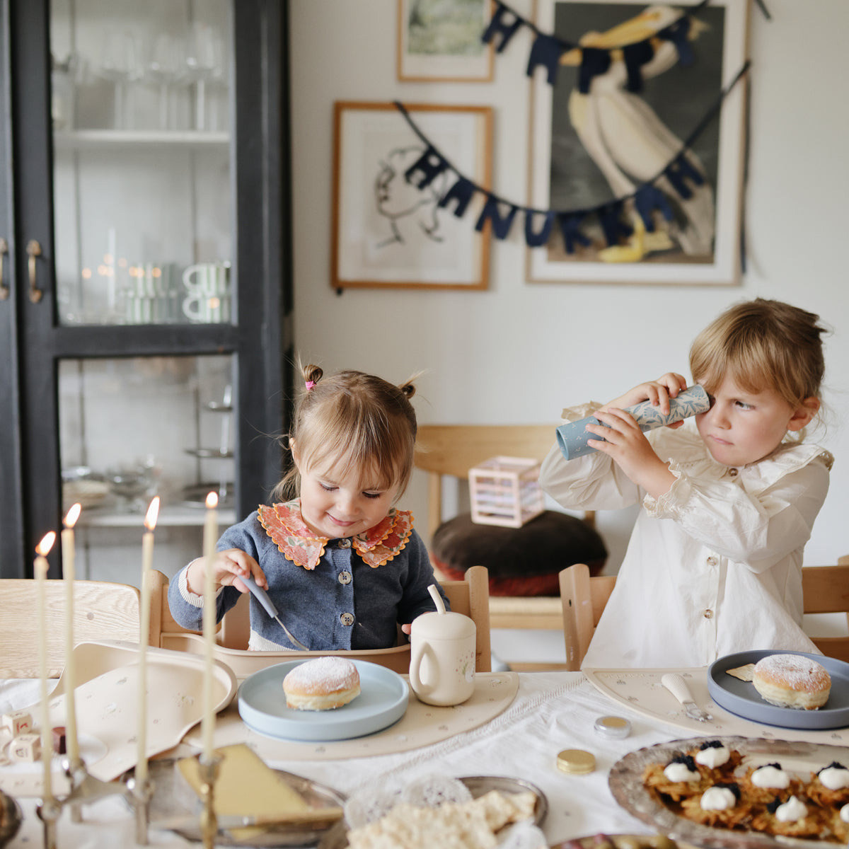 Two kids at Hanukkah; one eats, one uses a Mushie Classic Silicone Suction Plate.