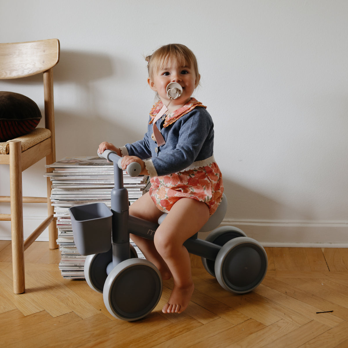 Toddler with FRIGG Rope Silicone Pacifier sits on gray tricycle indoors by magazines.