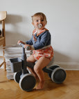 Toddler with FRIGG Rope Silicone Pacifier sits on gray tricycle indoors by magazines.