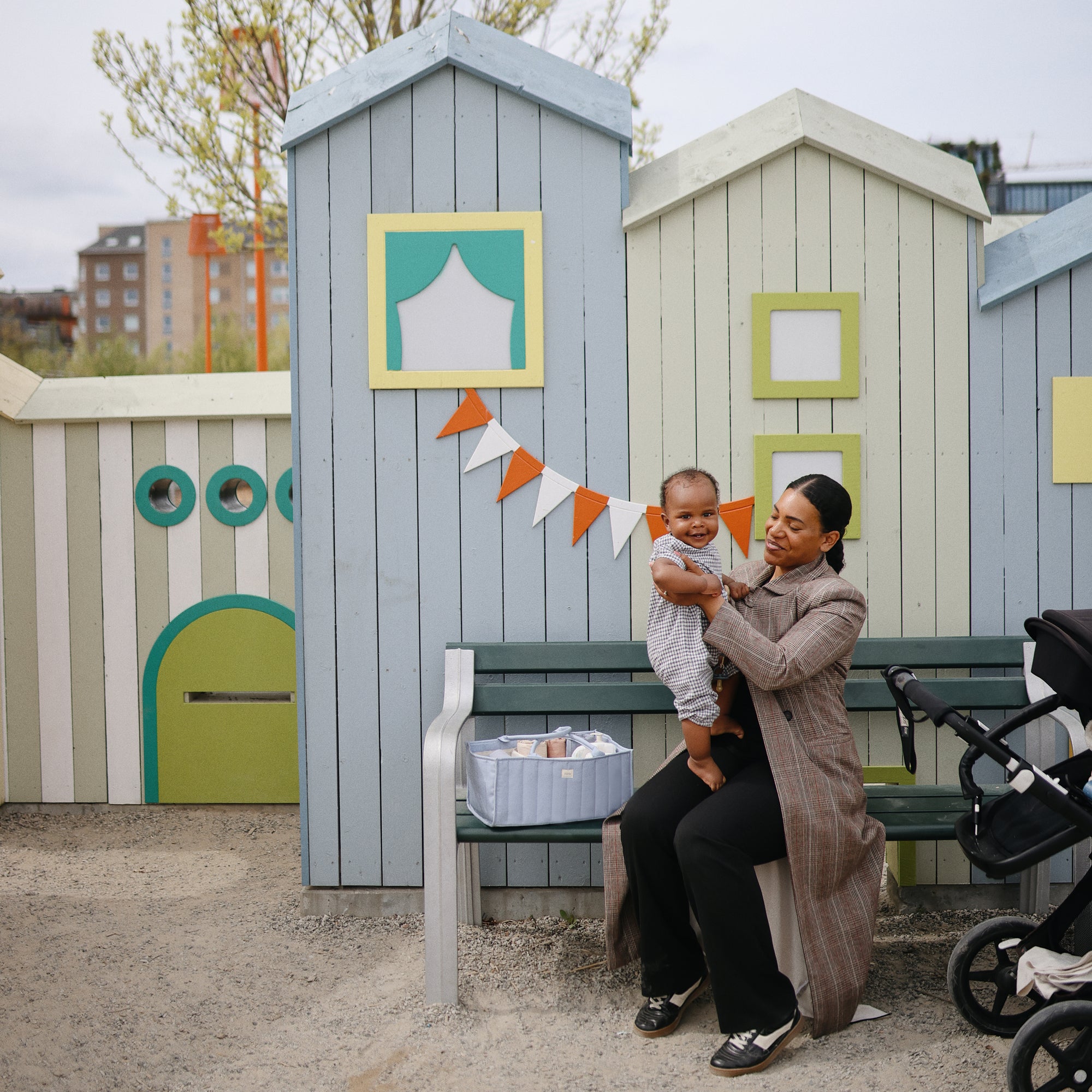 A woman sits with a toddler beside a mushie Diaper Caddy on a bench.