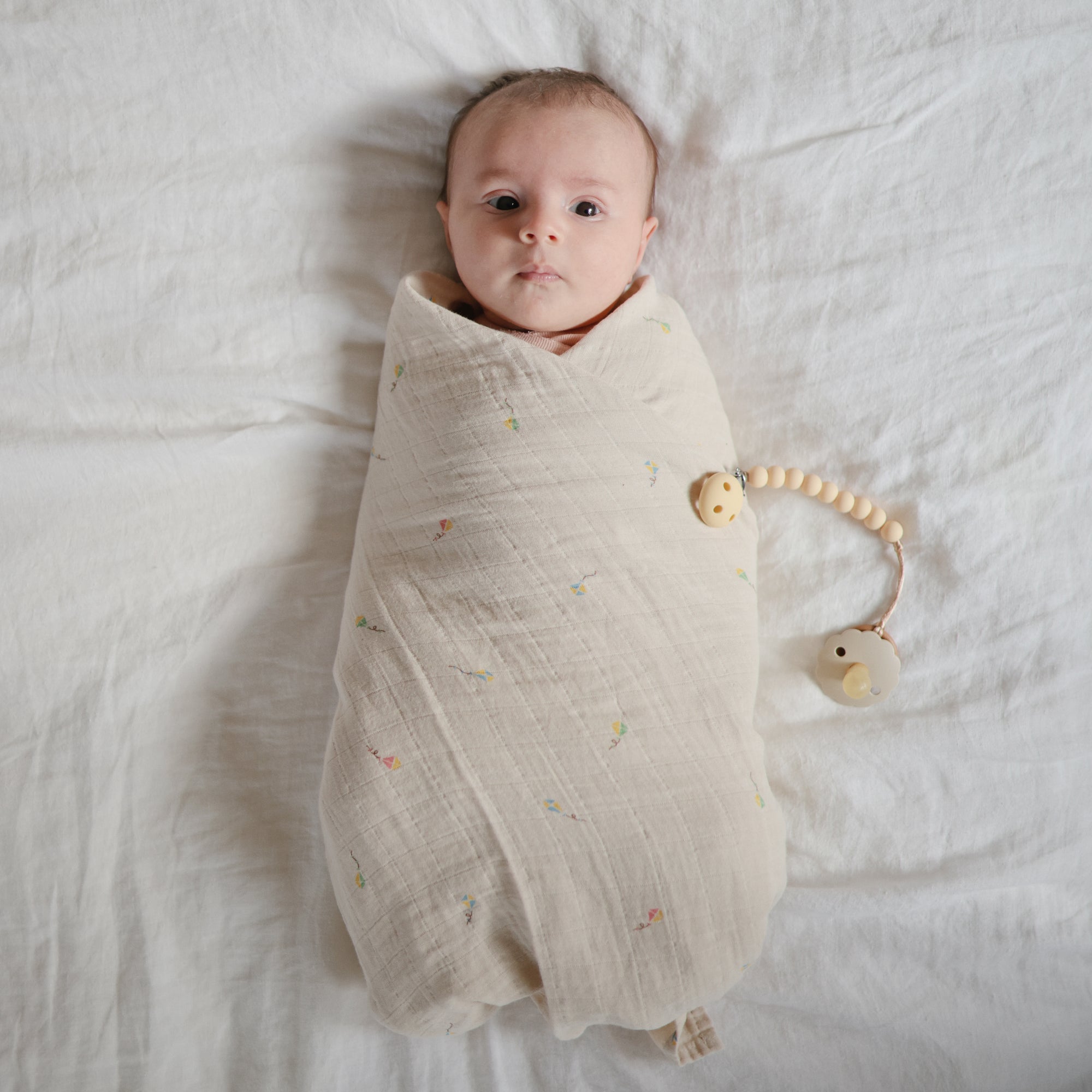 A baby wrapped in a mushie Organic Cotton Muslin Swaddle Blanket lies beside a pacifier clip.