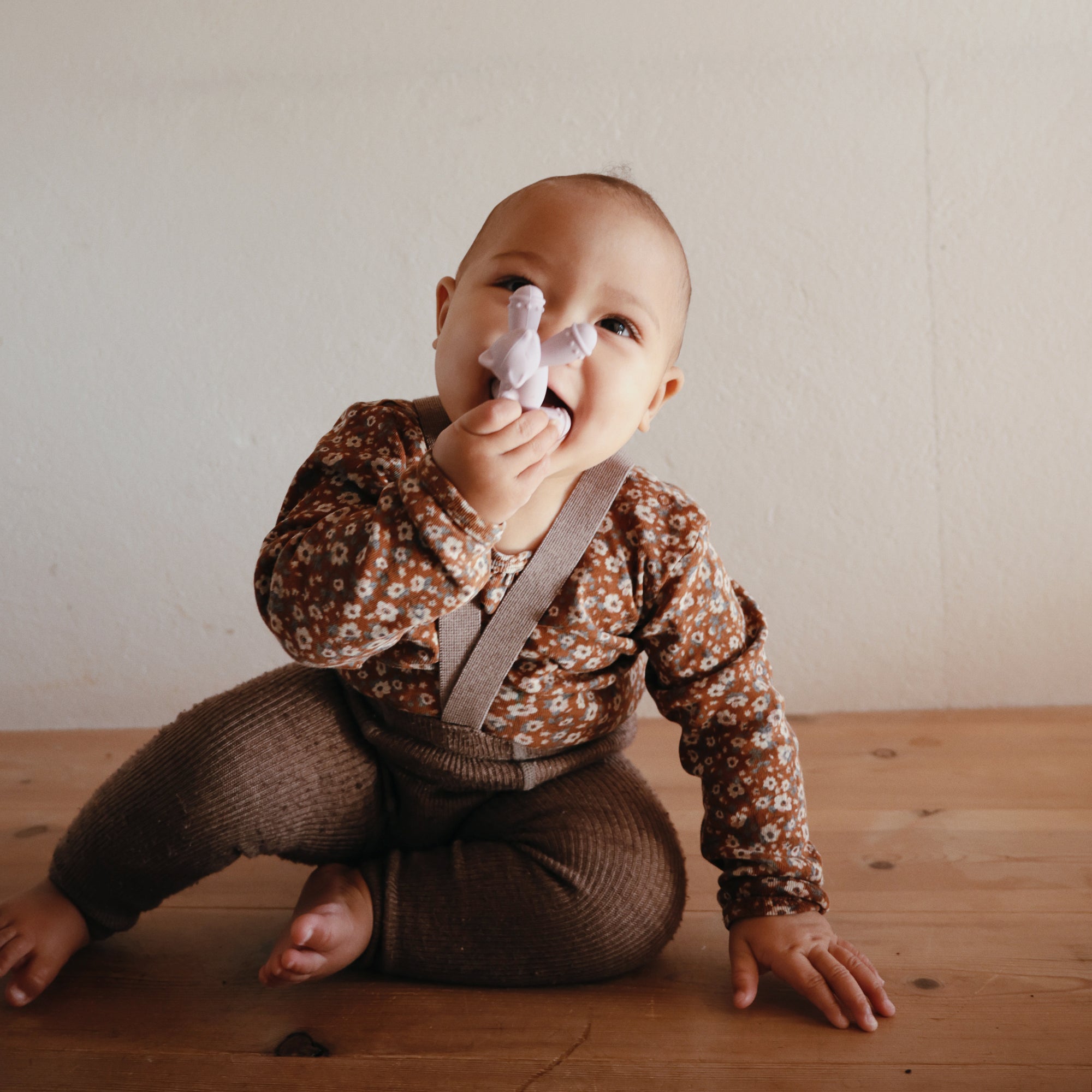 Smiling baby holds mushie Figurine Teether on wooden floor, in floral top and brown pants.
