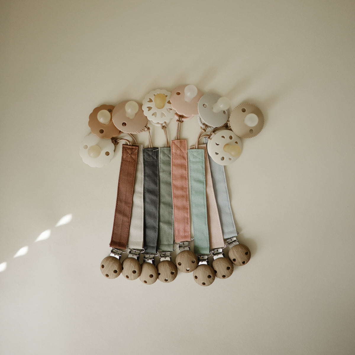 Mushie Linen Pacifier Clip 2-Pack: pastel linen clips with wood &amp; silicone, on cream surface.
