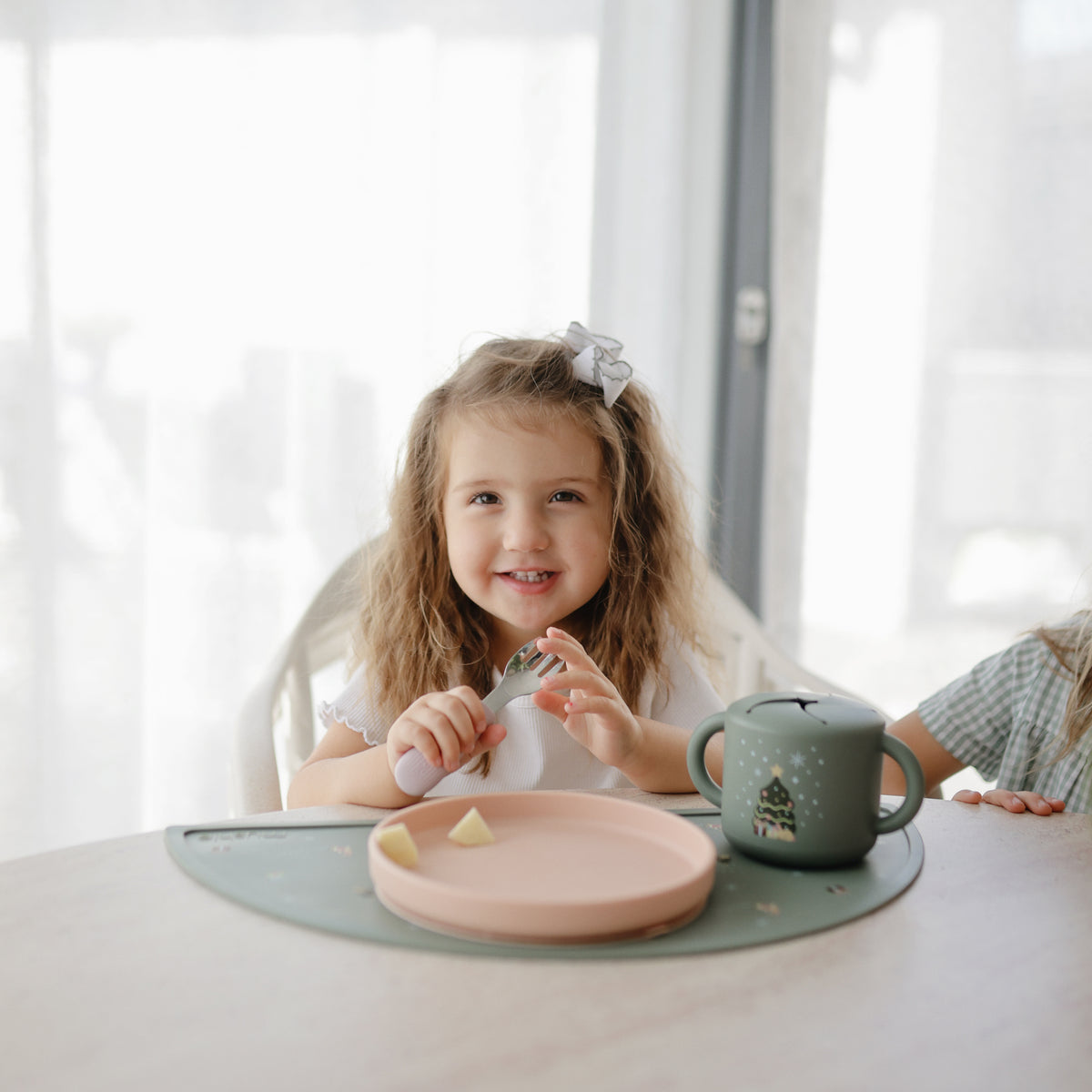Girl with a bow sits at table with food on mushie Silicone Placemat and mushie cup.