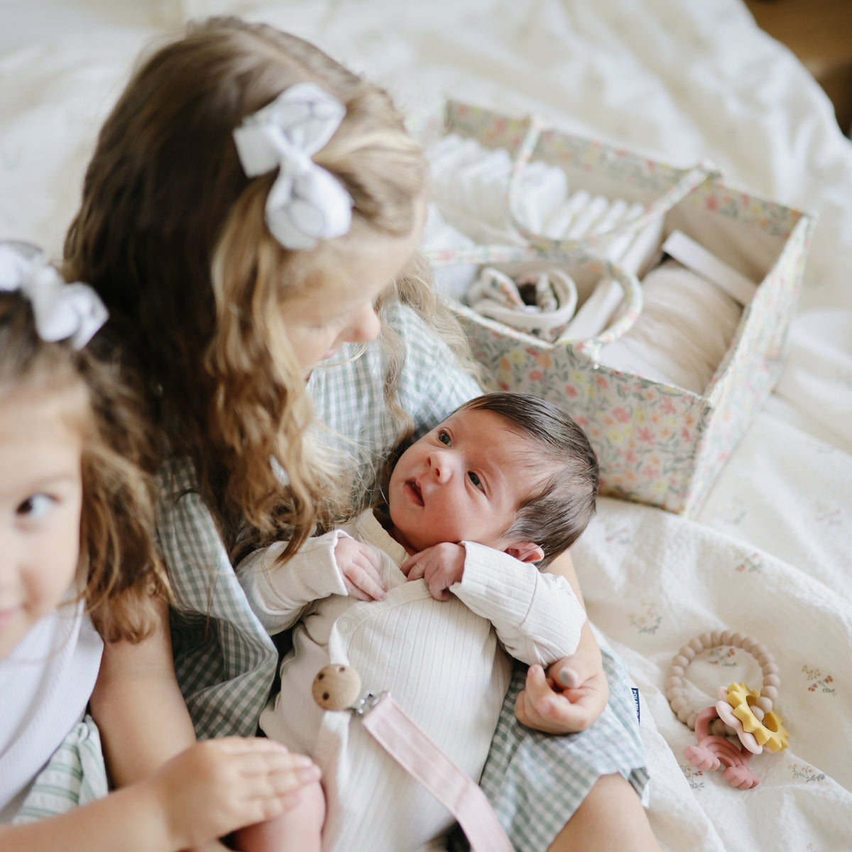 Young girl with bow holds newborn; mushie Teething Ring sits next to them on the bed.