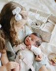 Young girl with bow holds newborn; mushie Teething Ring sits next to them on the bed.