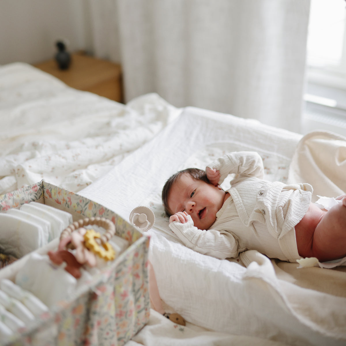 Newborn on bed next to mushie Extra Soft Muslin Changing Pad Cover and baby supply box.
