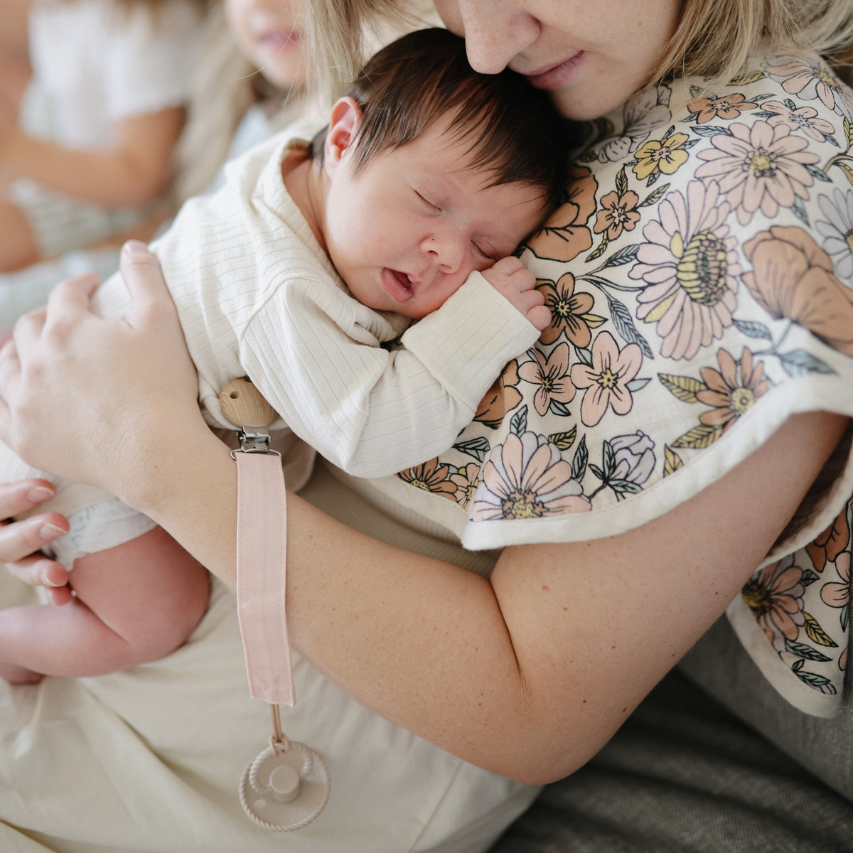 Baby with FRIGG Rope Silicone Pacifier rests on woman in a floral shirt, gently held.