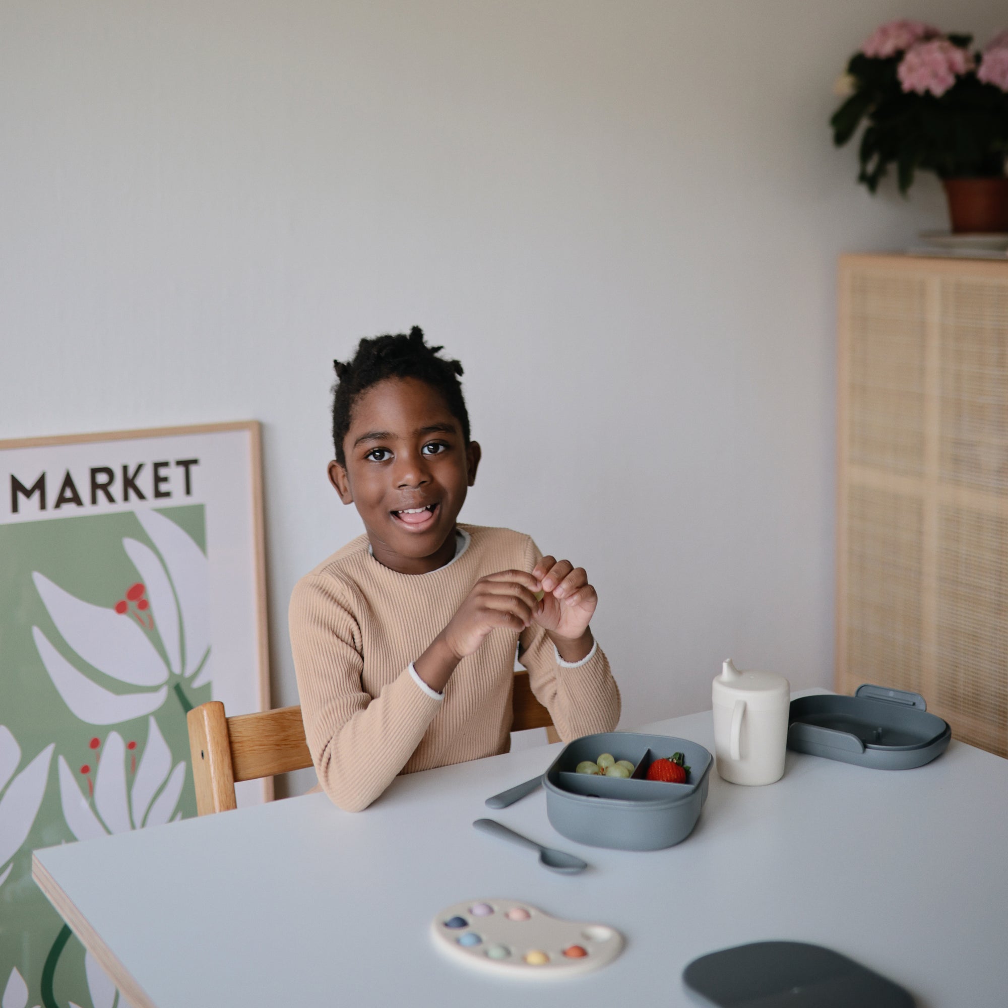 A smiling child enjoys fruit from a mushie Lunchbox with built-in compartments.