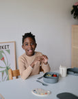 A smiling child enjoys fruit from a mushie Lunchbox with built-in compartments.