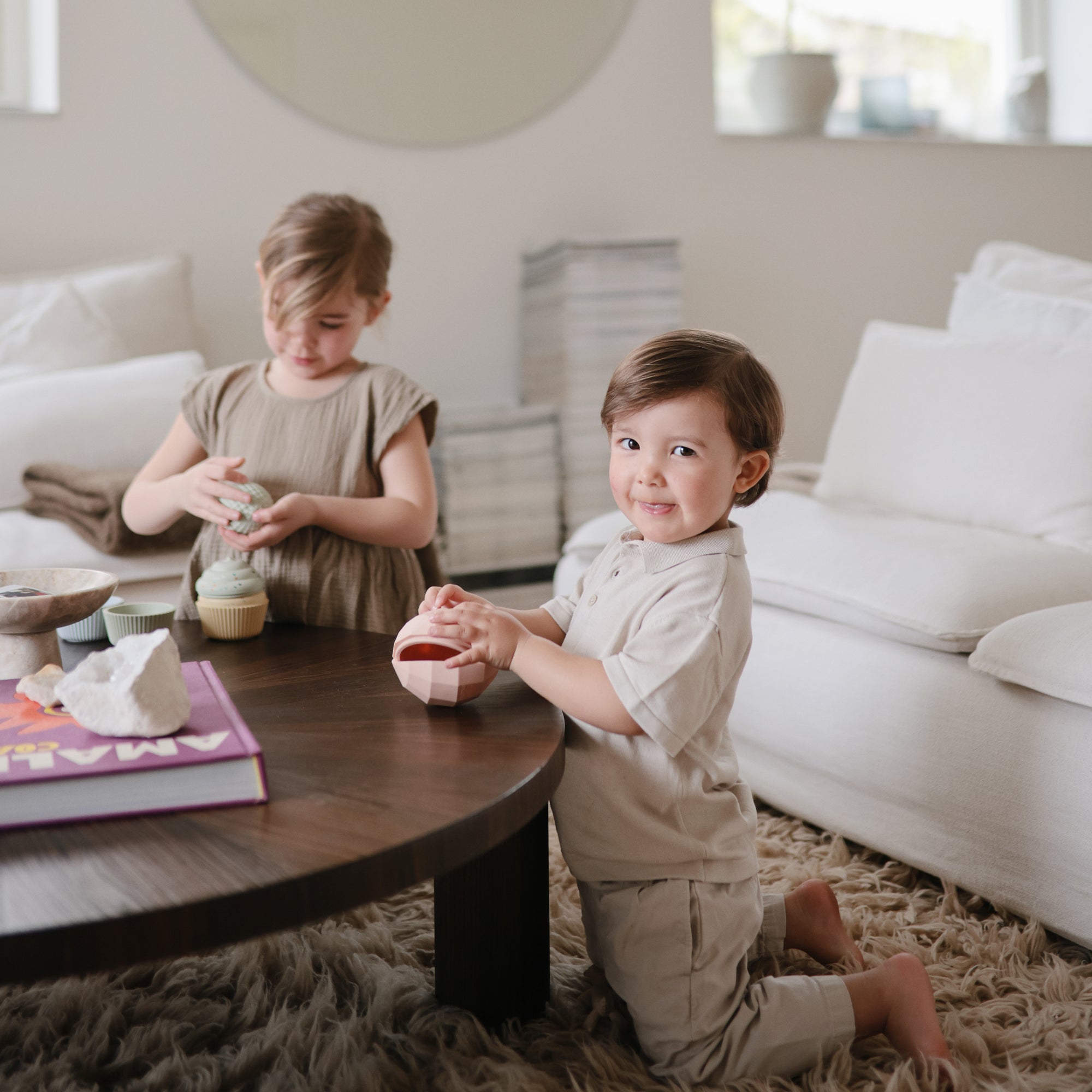 Two young children play with a mushie Nesting Spheres Sensory Toy on a coffee table.