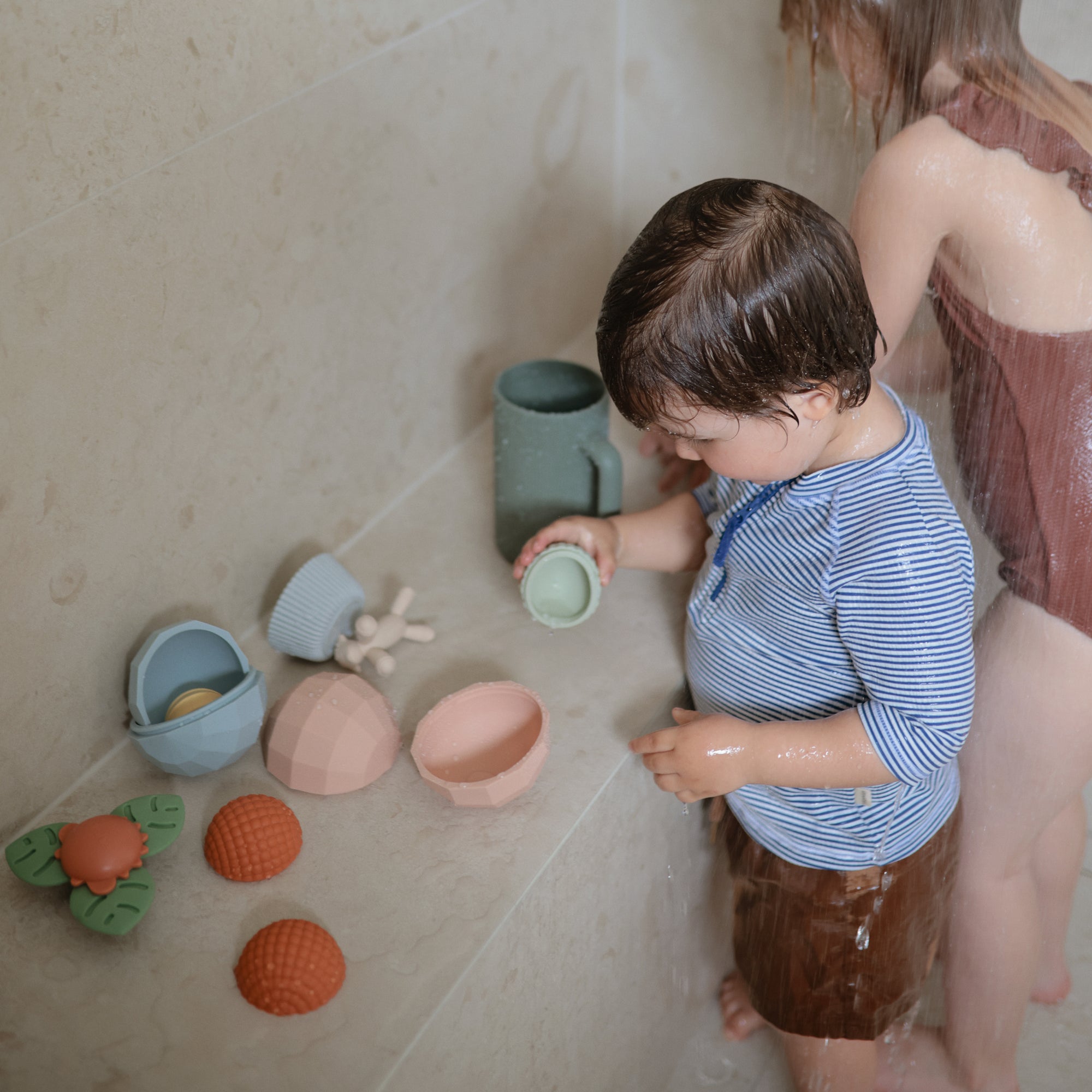 Two young children play with mushie Nesting Spheres Sensory Toy in pastel colors in a shower.