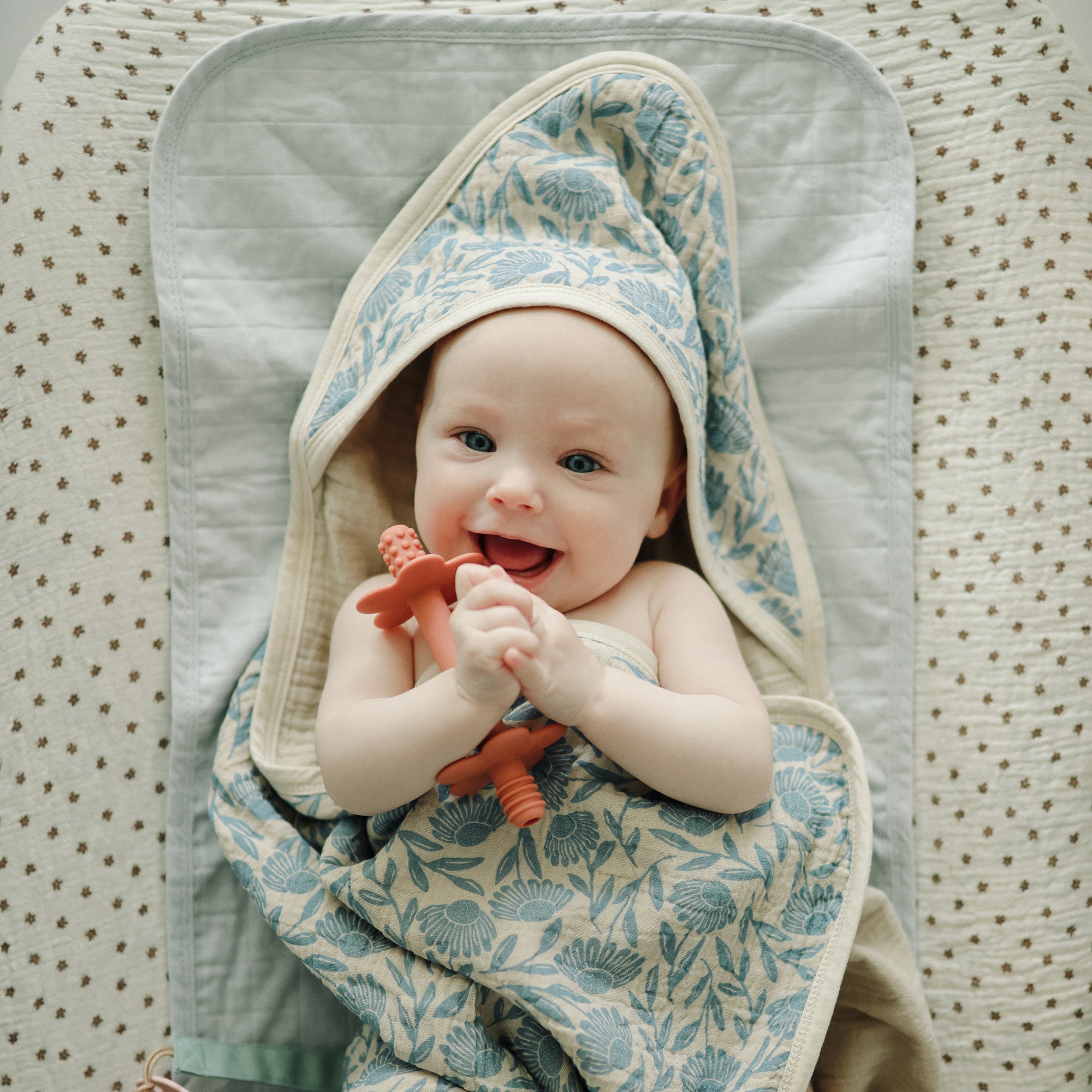 Smiling baby on Mushie Changing Pad Liners, wrapped in floral towel and holding a teether.