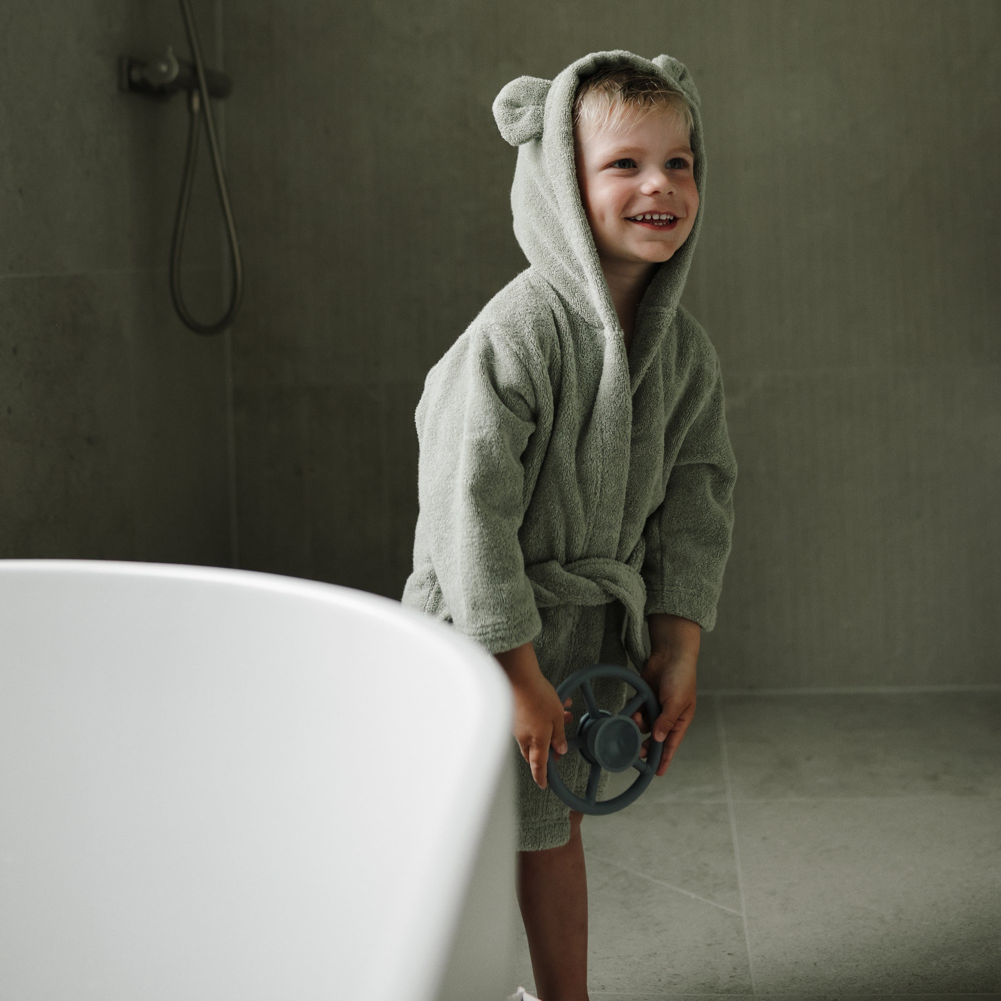 Smiling child in a mushie Bear Robe stands by a bathtub, holding a toy.