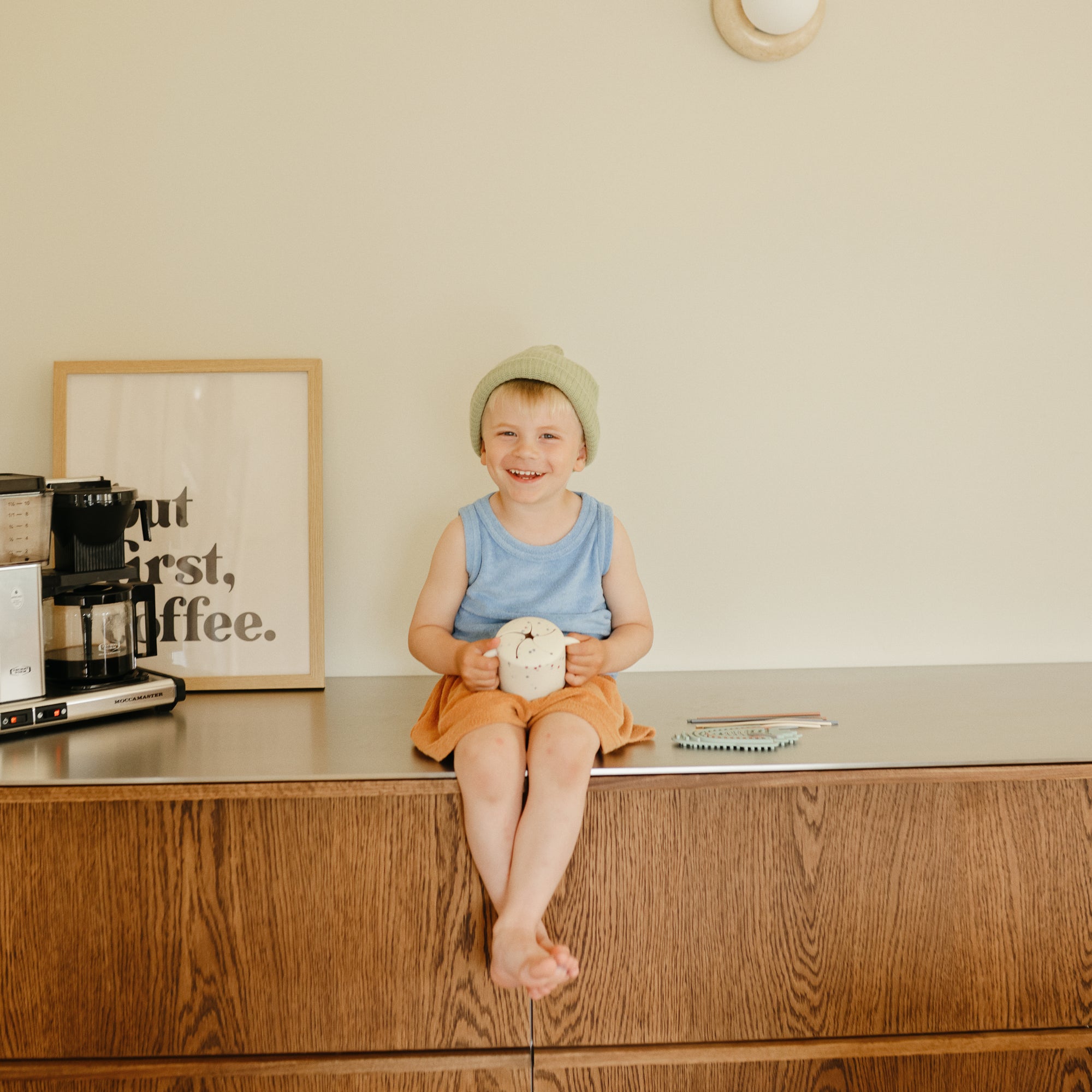Smiling child in beanie holds mug and mushie Silicone Sensory Toy, coffee sign behind.