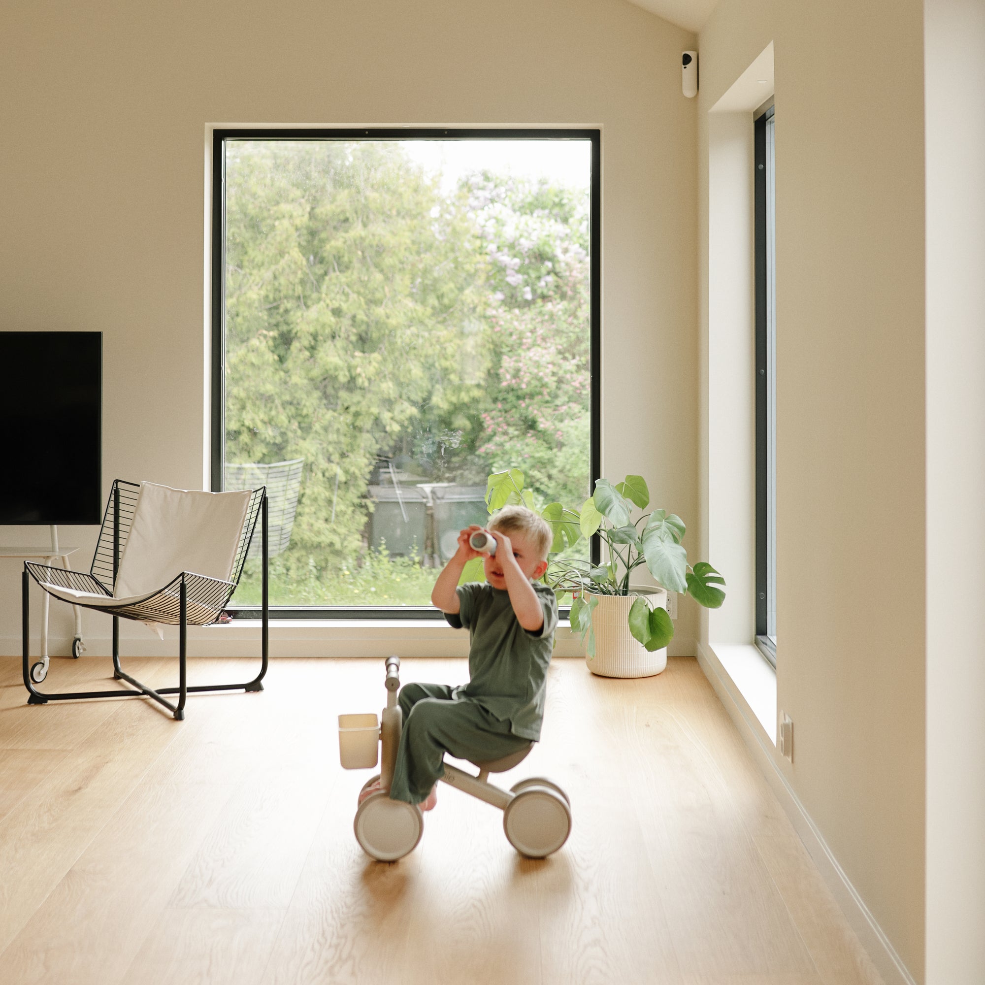 A young child rides a mushie Mini Toddler Bike, scanning the room with binoculars.