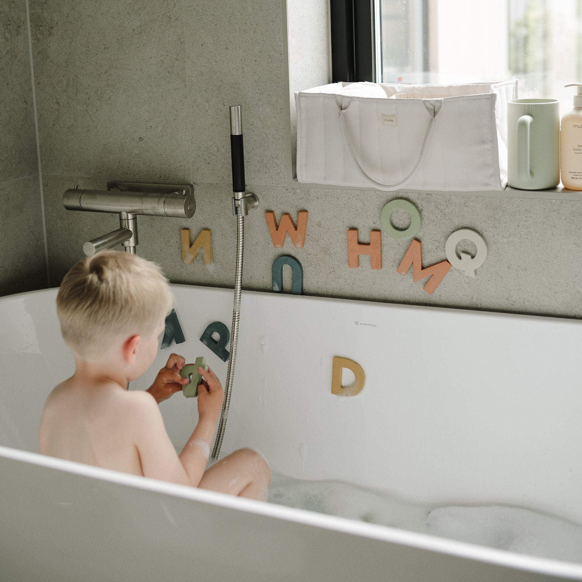 A mushie Diaper Caddy sits by the window as a young child plays with foam letters in the tub.