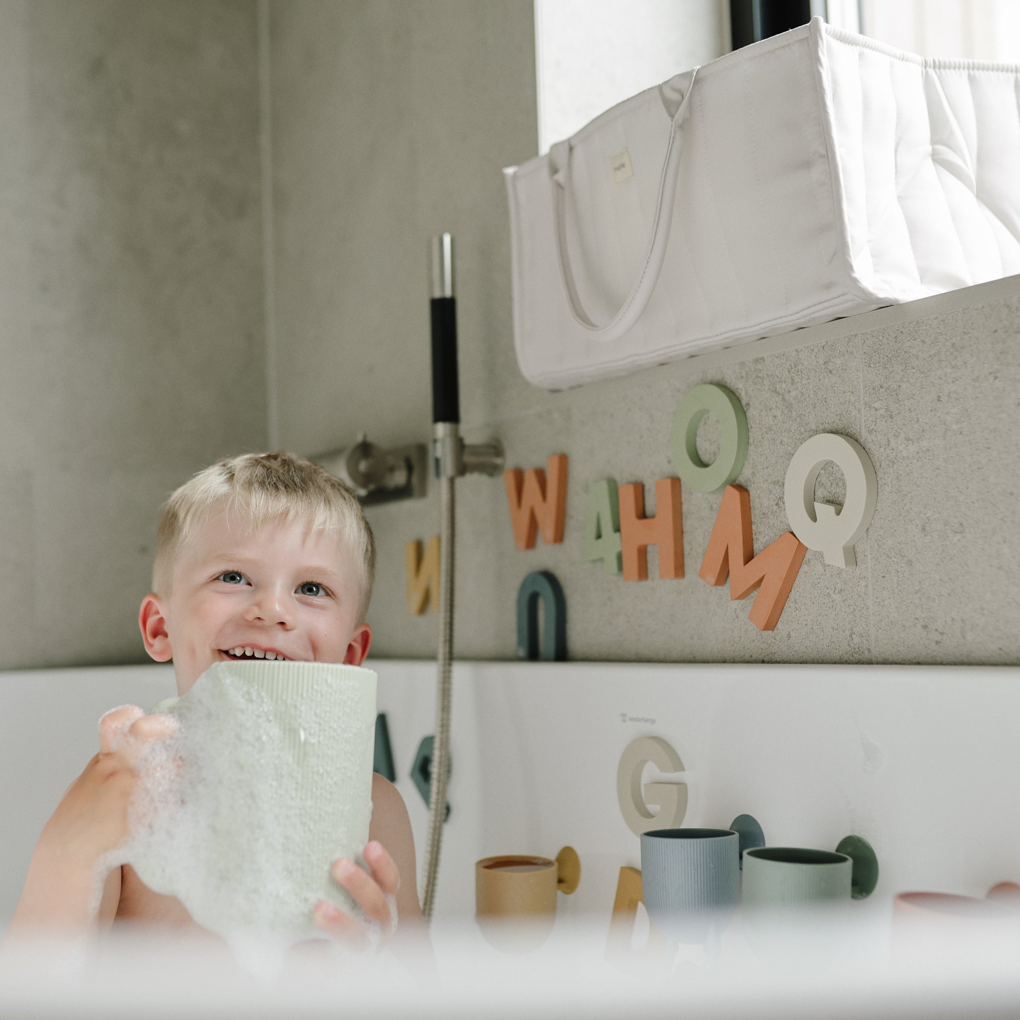 Smiling child enjoys bath with bubbles, colorful letters, and a Mushie Bath Rinse Cup nearby.