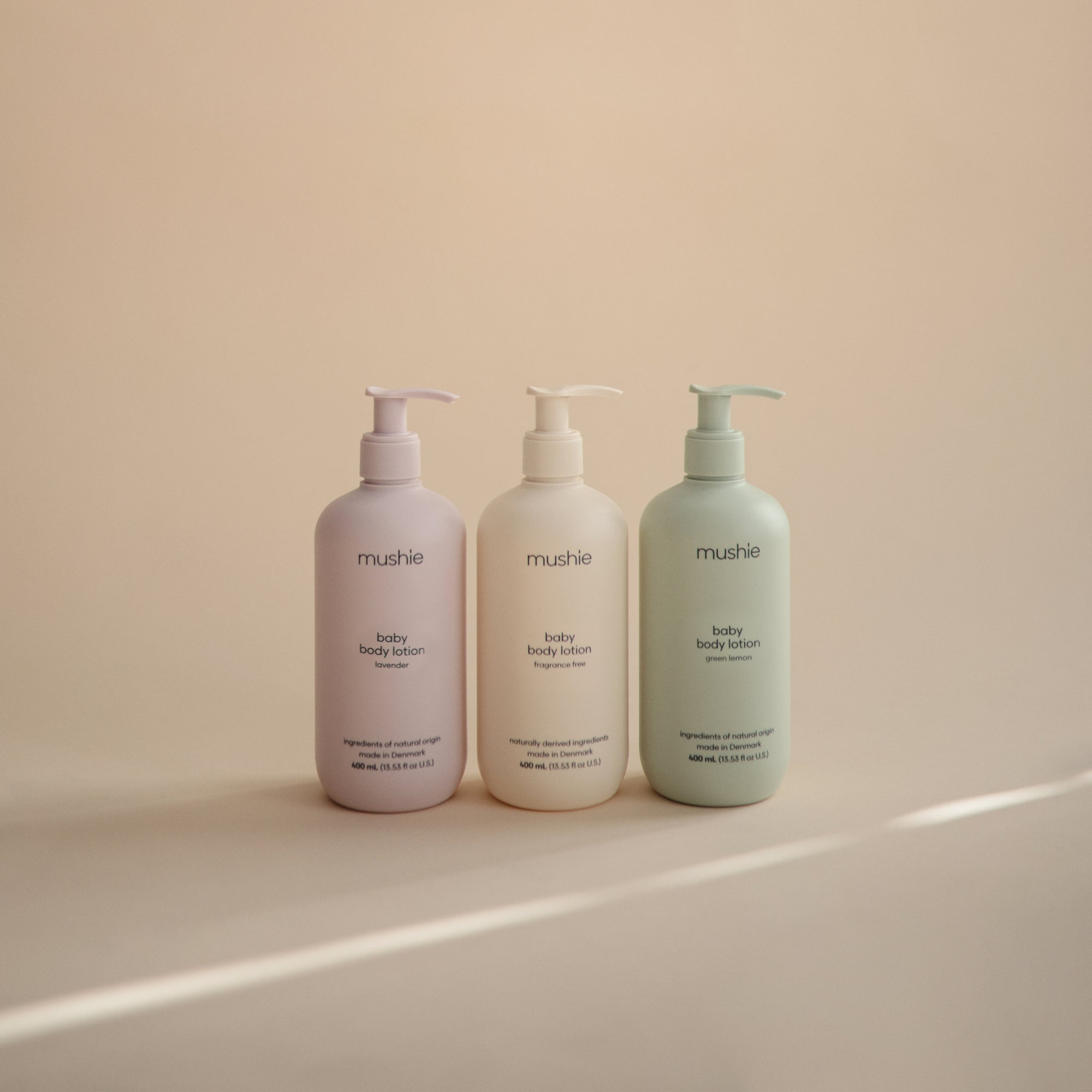 Three mushie Baby Body Lotions in pastel bottles with natural ingredients on a neutral background.