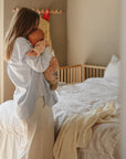 Woman wraps baby in mushie Organic Cotton Muslin Swaddle Blanket beside a cozy bed.