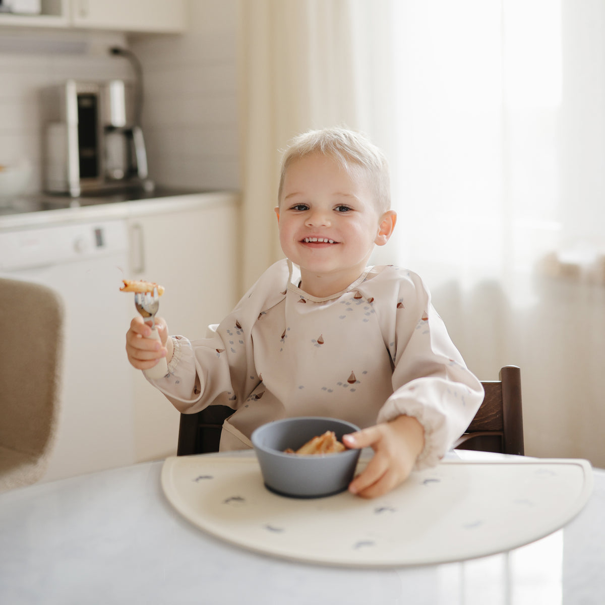 Smiling toddler wearing a mushie Long Sleeve Bib while eating at a table.